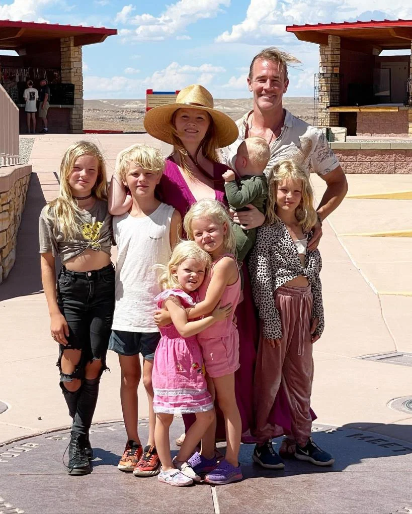 James Van Der Beek, his wife Kimberly, and their six children pose outdoors in front of a building with hills in the background.