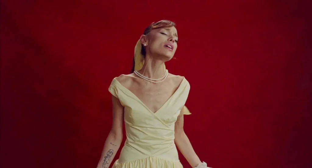 Ariana Grande singing in a yellow dress, with a yellow scarf and pearl necklace.
