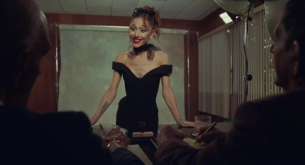 Ariana Grande as Penny Pingleton from "Hairspray" in a black dress, smiling while standing at a table before two men.
