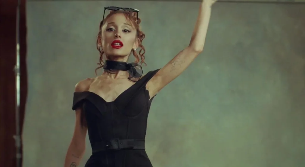Ariana Grande in a black off-the-shoulder dress with red lipstick, black neck scarf, and sunglasses on her head, with one arm raised.