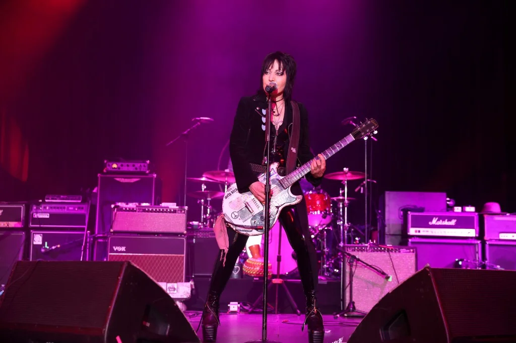 Joan Jett performing on stage, playing a white electric guitar with stickers, dressed in a black leather jacket and black shiny pants.