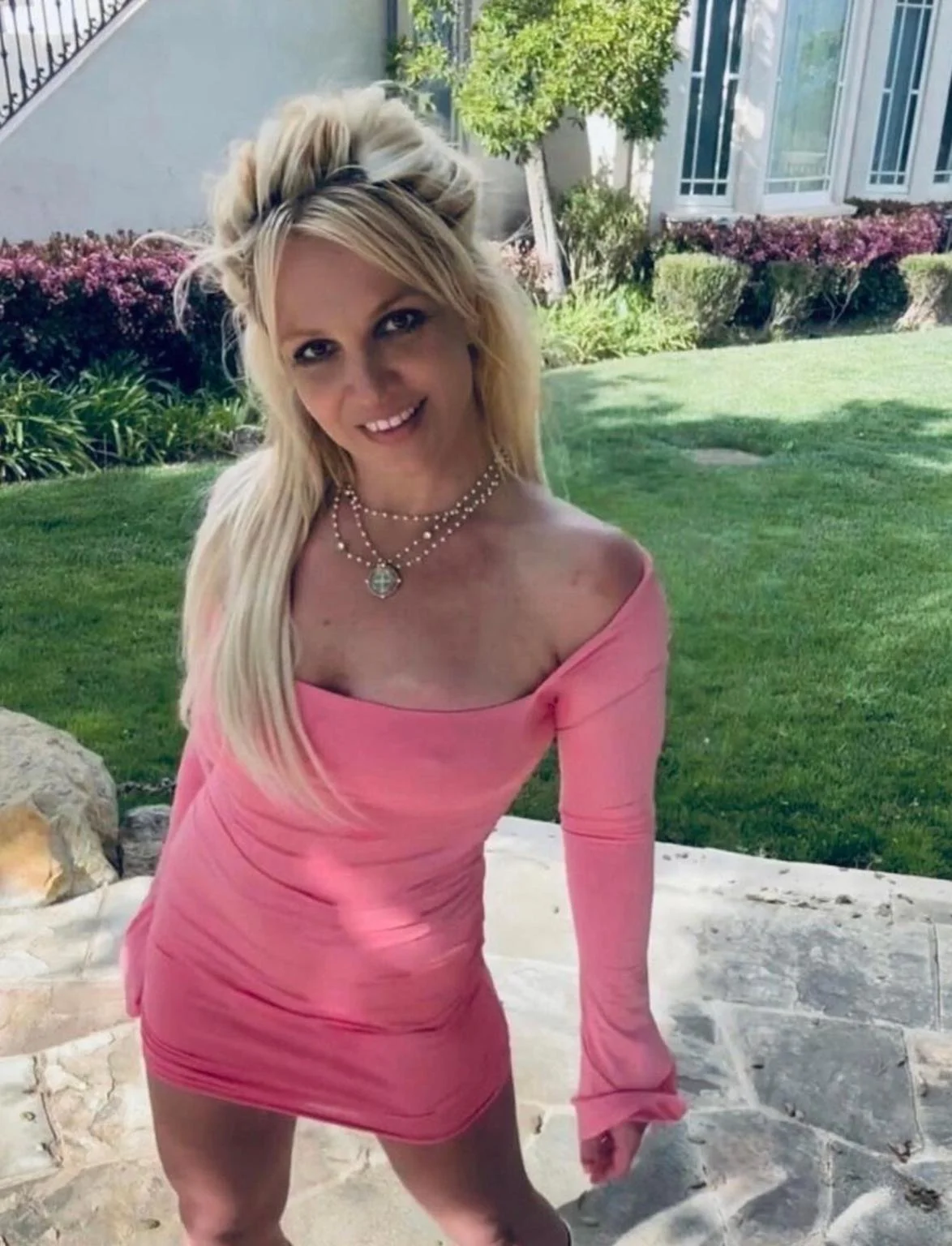 Britney Spears posing in a pink dress in front of a house and garden.