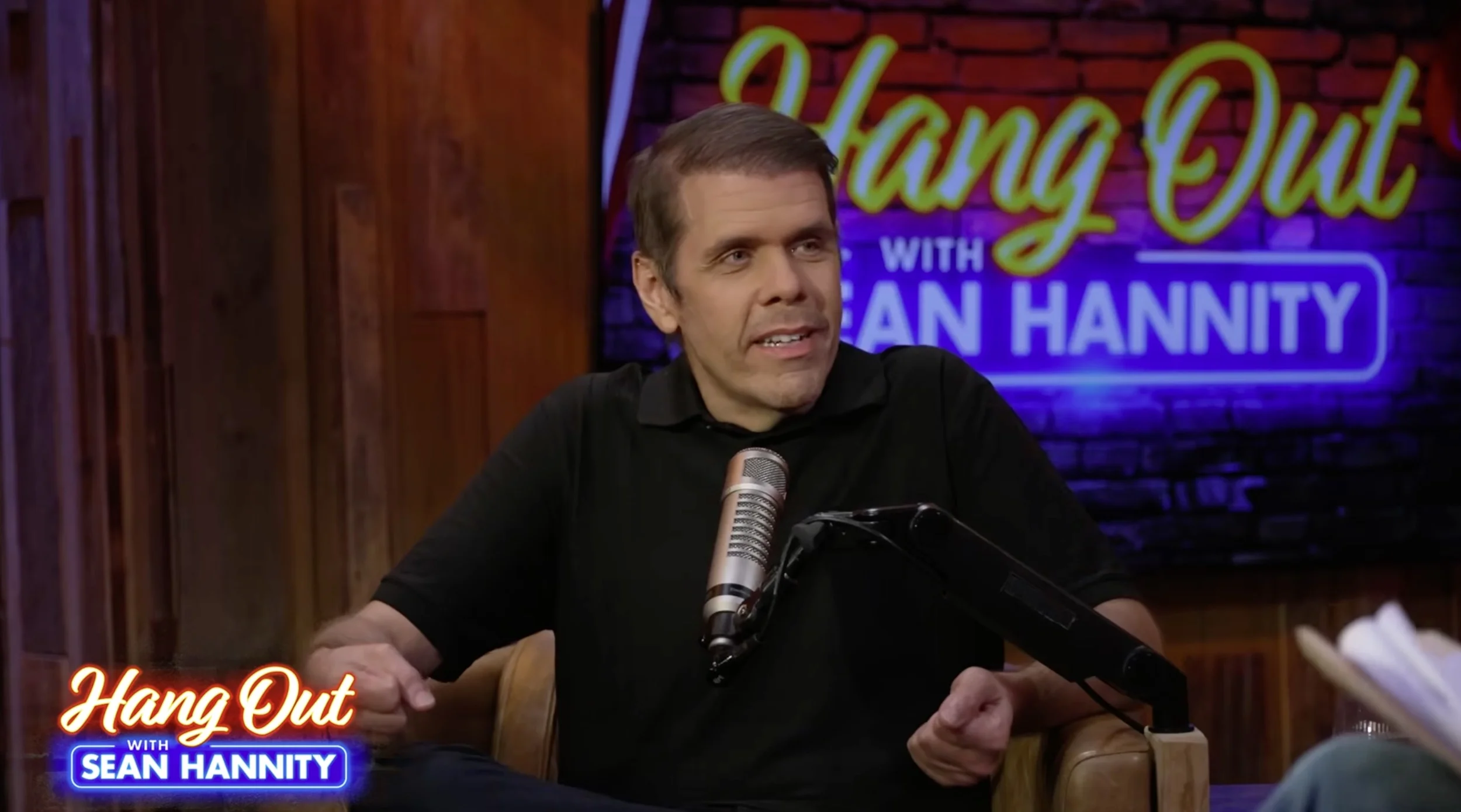 Perez Hilton wearing a black polo shirt while speaking into a microphone.