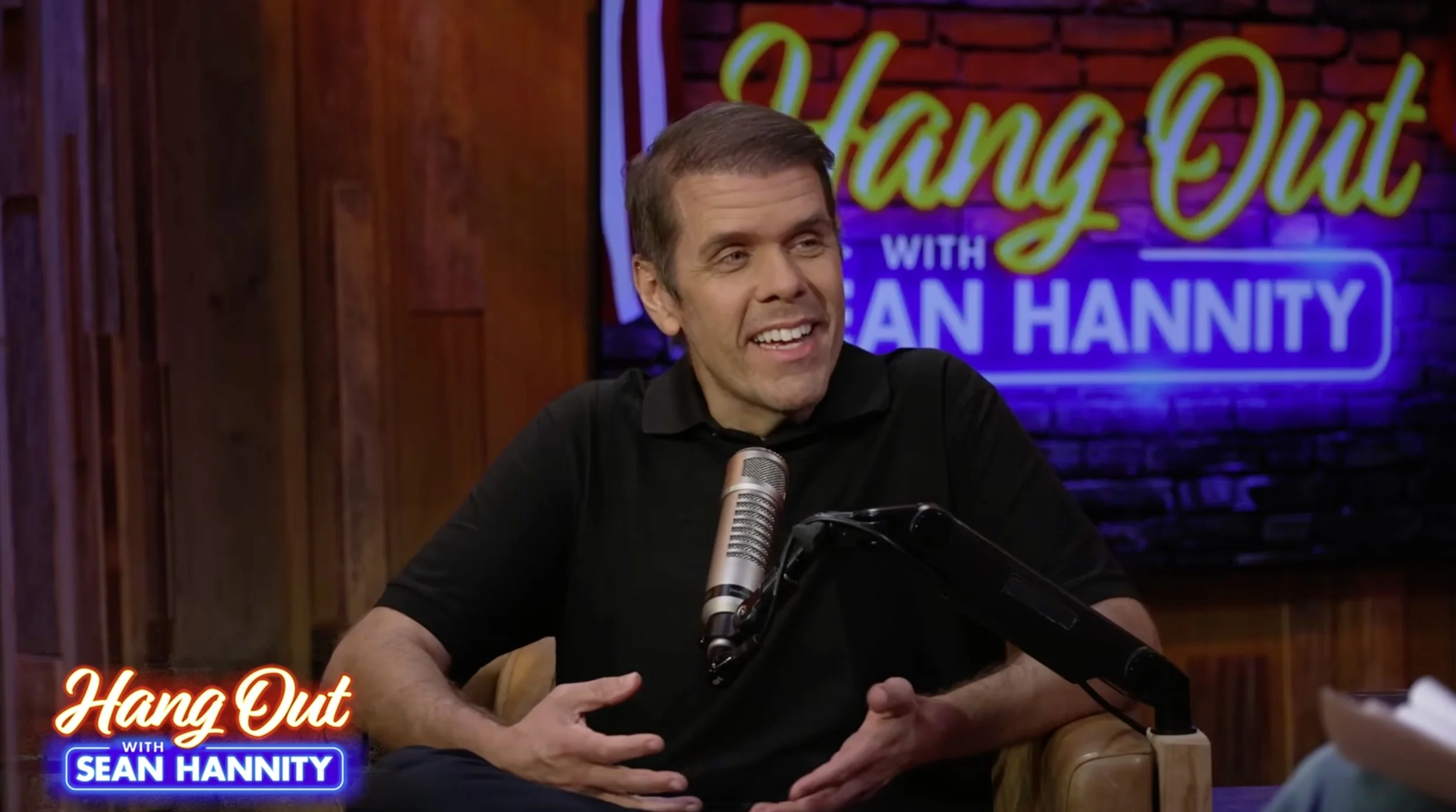 Perez Hilton sitting on a talk show set with a microphone in front of him, gesturing with his hands.