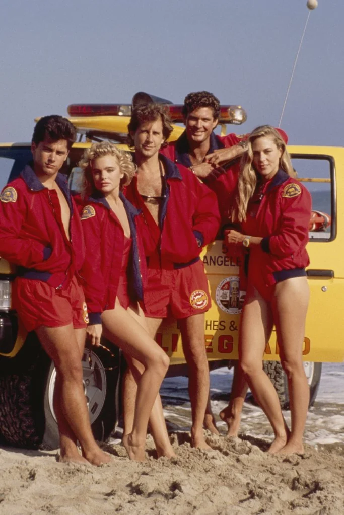 Billy Warlock, Erika Eleniak, Parker Stevenson, David Hasselhoff, and Shawn Weatherly pose on a beach with a lifeguard vehicle.