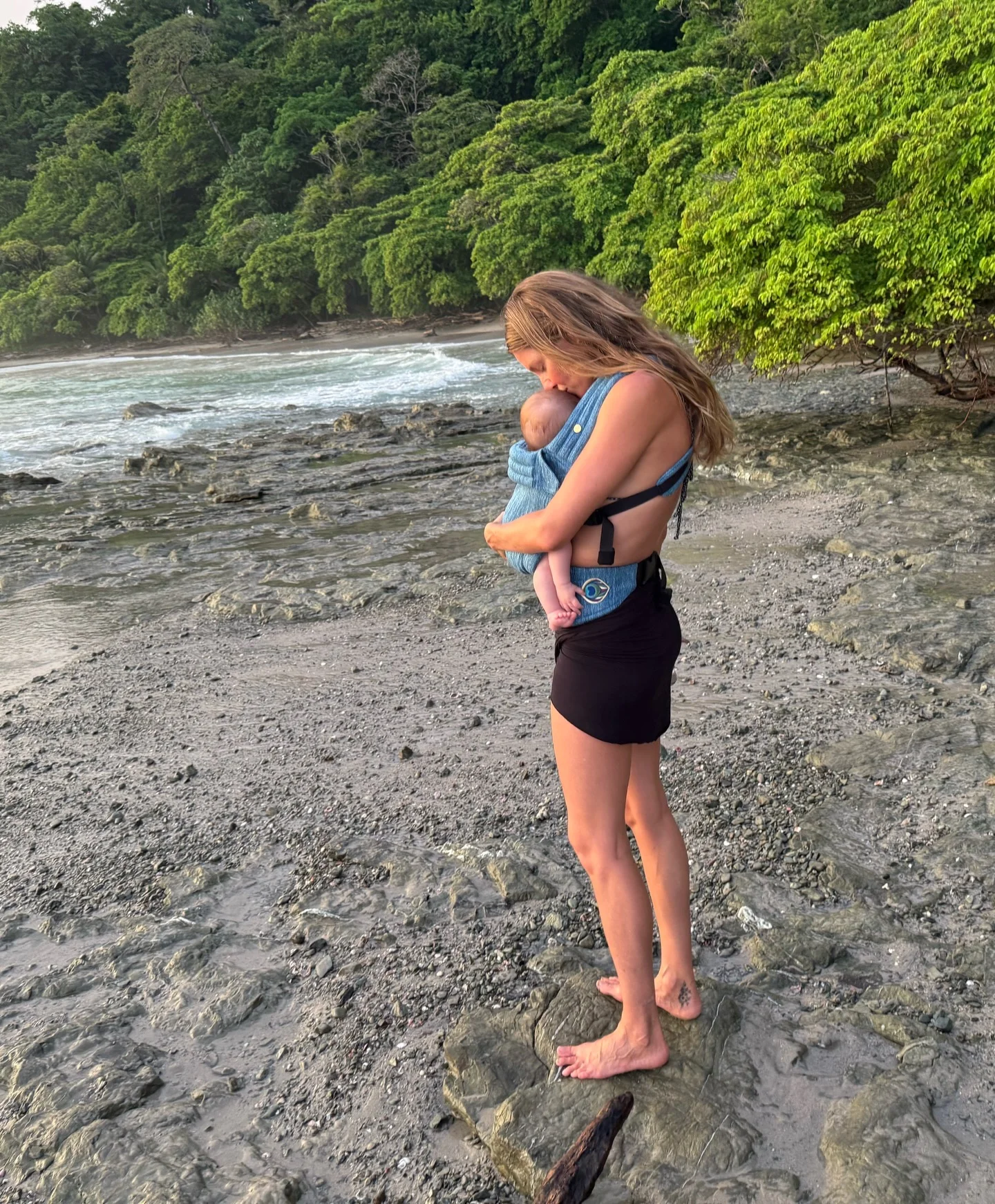 Gisele Bündchen holding her baby in a carrier on a rocky beach.