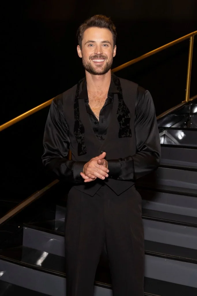 Dylan Efron smiling in a black shirt, vest, and trousers with a gold railing and stairs in the background.