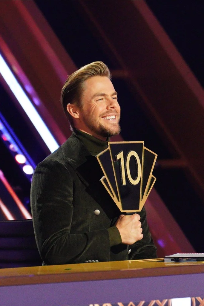 Derek Hough holding up a score paddle with the number 10.
