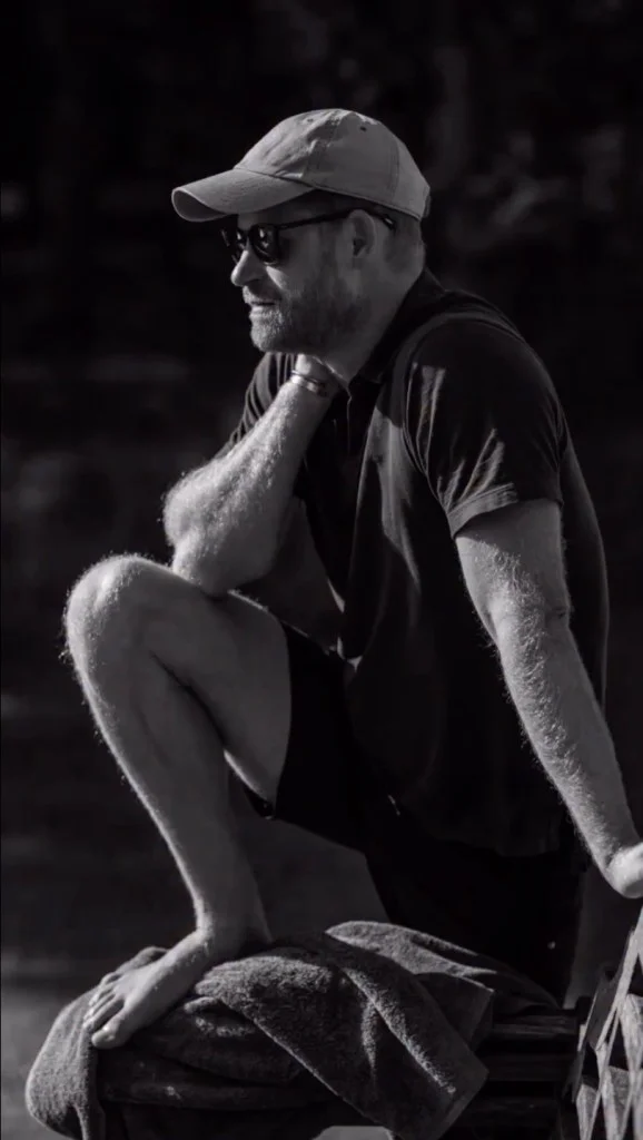 Prince Harry in a baseball cap, sunglasses, t-shirt, and shorts, crouching with one foot on a towel.
