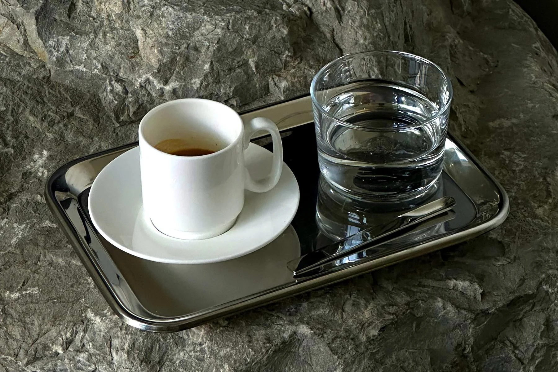 a close-up imagine showing a a steel tray holding a small glass of water and a white cup and saucer of espresso, sitting on top of a rock 