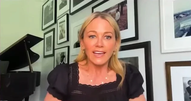 Christine Taylor speaks into the camera, with a black piano and framed pictures on the wall behind her.