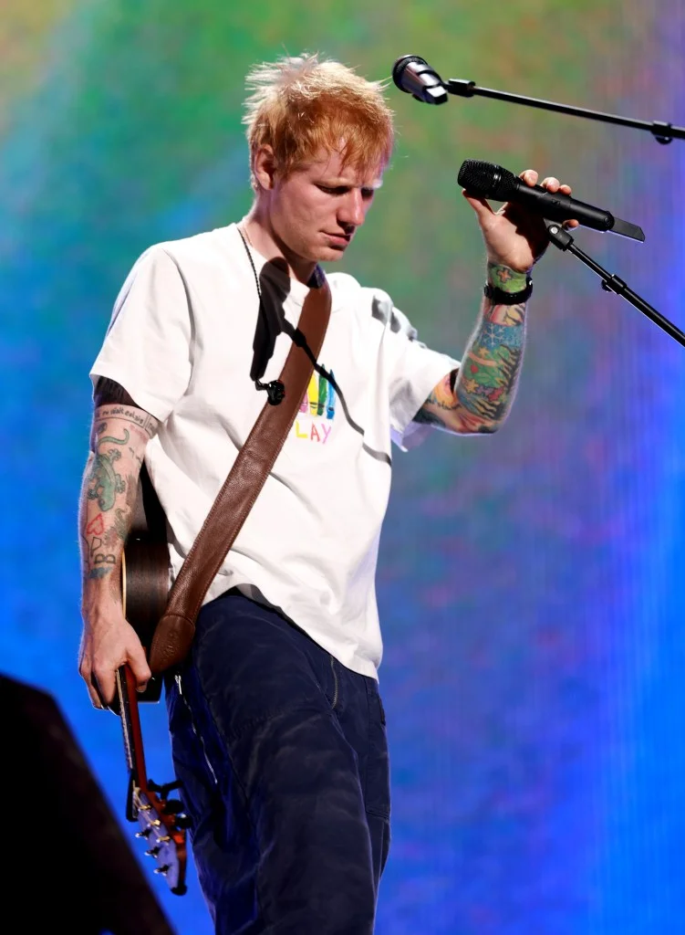 Ed Sheeran performing onstage, holding a microphone in his right hand and an acoustic guitar in his left.