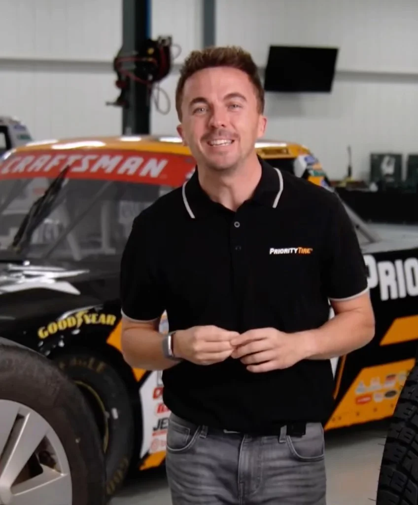 Actor Frankie Muniz stands in a garage with a race car behind him.