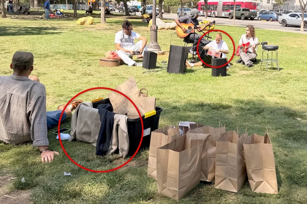 Three musicians performing in a park with a box of merchandise and several paper bags.