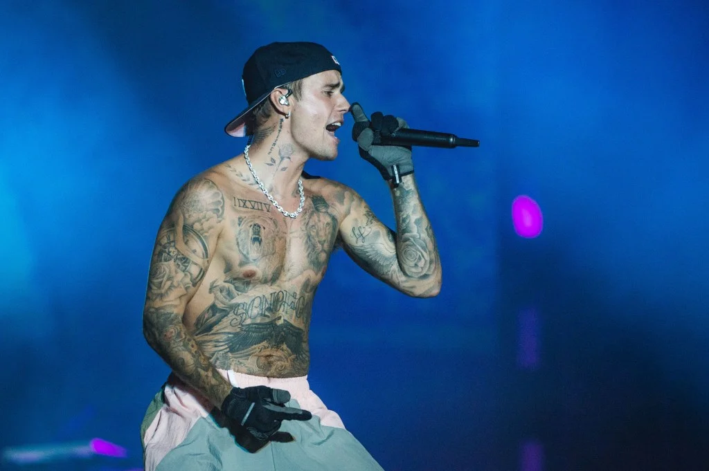 Justin Bieber performing live, shirtless, wearing a black cap, chain, and black gloves, holding a microphone.