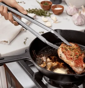 hand holding silver tongs gripping meat in a cast-iron skillet on a stove.